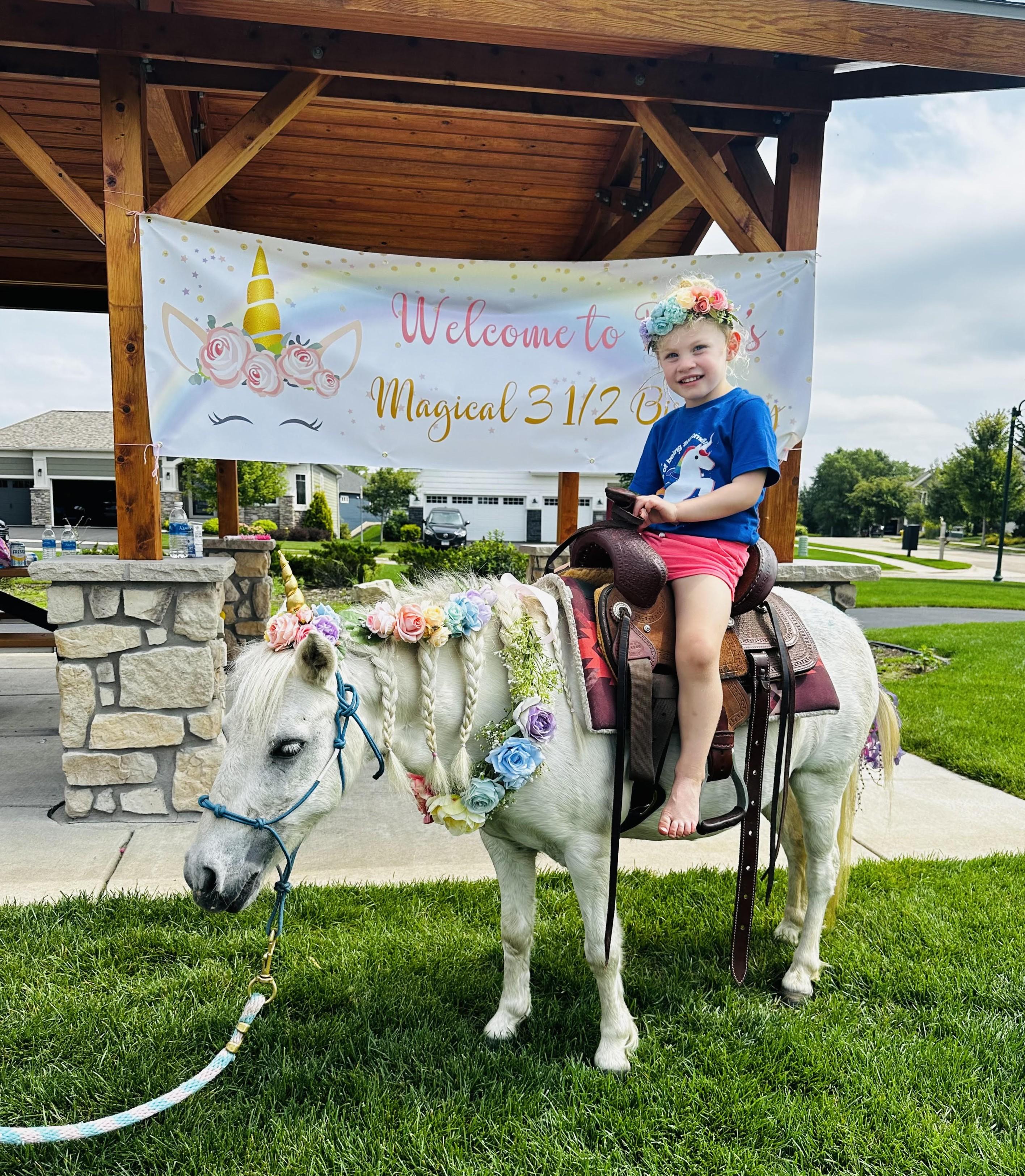 A young girl in a flower crown sitting in the saddle on a white pony decorated with a braided, flower-woven mane at a backyard birthday party