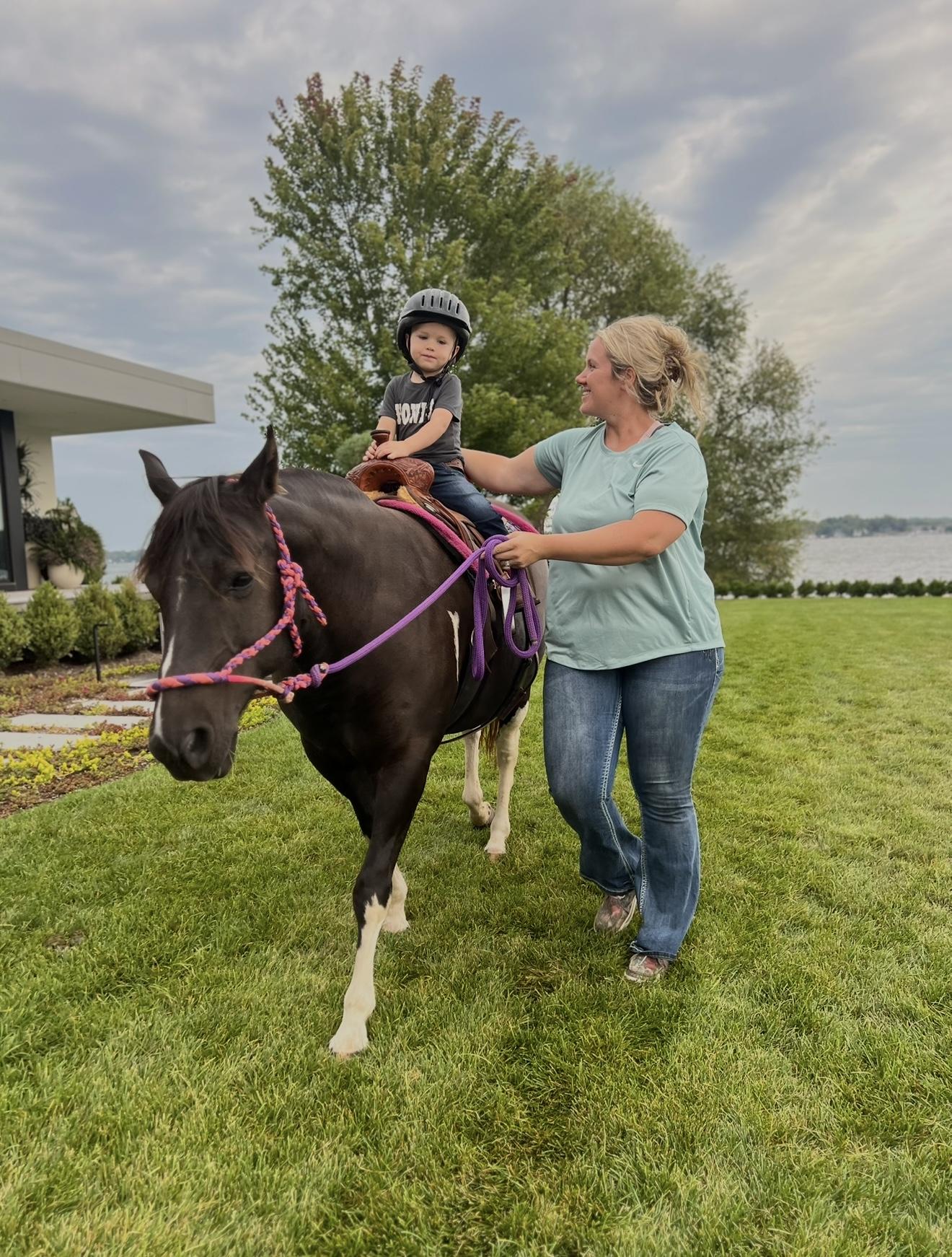 A toddler on a first pony ride, led by a handler across a backyard