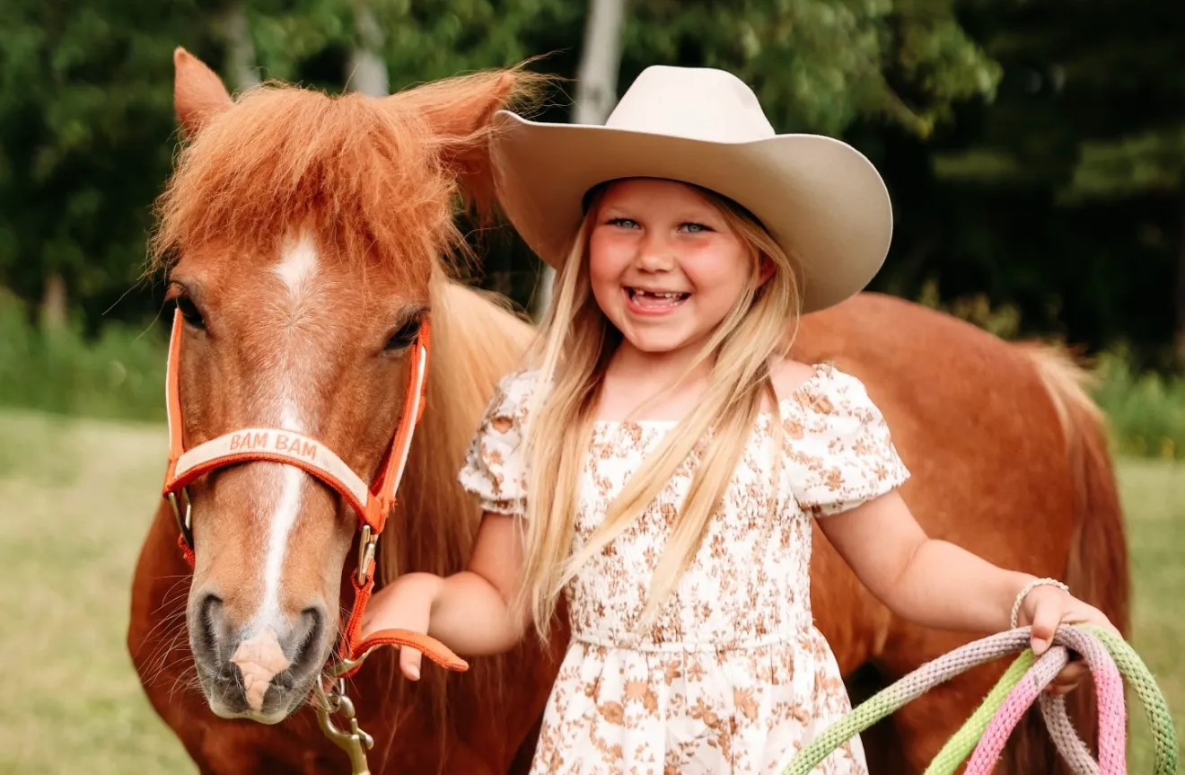 A young cowgirl in a cowboy hat standing with Bam Bam the pony