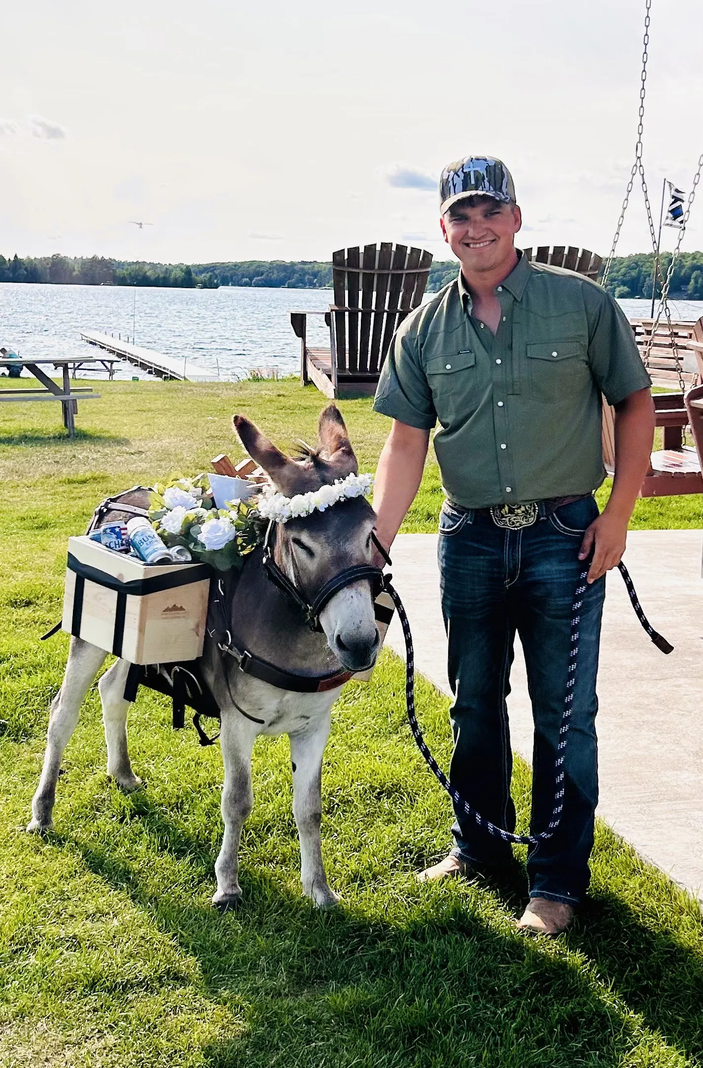 Kyle Knops standing lakeside with Guapo, a grey miniature donkey wearing a white flower crown and a wooden saddle bar stocked with cold cans